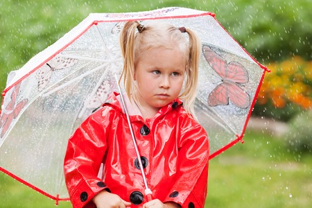 Serious pensive pretty little girl in red raincoat with umbrella walking in park summer, ladybug costume, portrait, rain, outdoorの写真素材