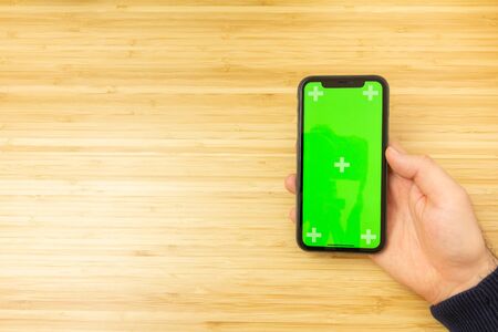 Mockup image of hand holding black mobile phone with green screen on the office table on the...