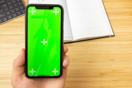Mockup image of hand holding black mobile phone with green screen on the office table on the background with stationery tools. San Francisco, February 2020のeditorial素材