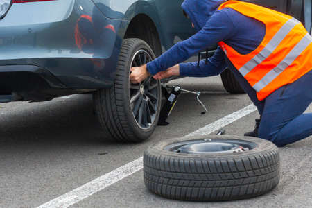 A man in a orange safety vest changes a flat tire on a road. Closeup mans hands to the wheel of a...