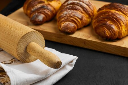 Close up of croissants on a wooden desk, a rolling pin and a kitchen towel on a black table....