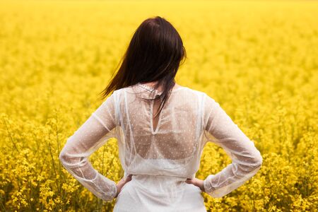 Woman in a white dress standing back in a yellow rapeseed field. Spring concept.の写真素材