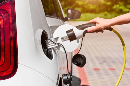 Close up woman hands plugging a power supply cable to electric car for charging at charging station outdoor.の写真素材