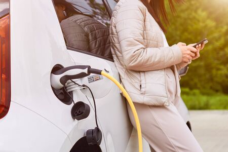Young woman charging an electric car at public charging station and pays using a mobile phone. Innovative eco-friendly vehicle.の写真素材