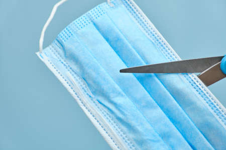 Scissors cutting a surgical face mask against coronavirus disease on a blue background. Cut mask...