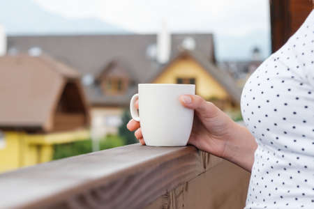 Close up womans hand holding a white cup with coffee or tea on the balcony with an amazing view on the mountains and villas roofsの写真素材