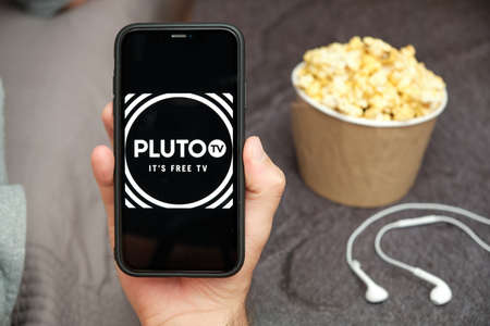 Close up mans hand holding a mobile phone with Pluto tv logo with Apple earphones and popcorn box next to him, Free TV concept, August 2020, San Francisco, USAのeditorial素材