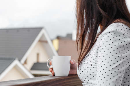 Young woman in polka dot T-shirt with a white cup of tea or coffee standing on the balcony. Morning in the countryside. Concept of relax.の写真素材