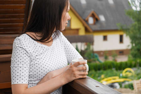 Young woman in polka dot T-shirt with a white cup of tea or coffee standing on the balcony. Morning in the countryside. Concept of relax.の写真素材