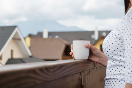 Close up a white cup in a woman hand on the wooden balcony on the background of mountains and house roof. Morning on the vacationの写真素材