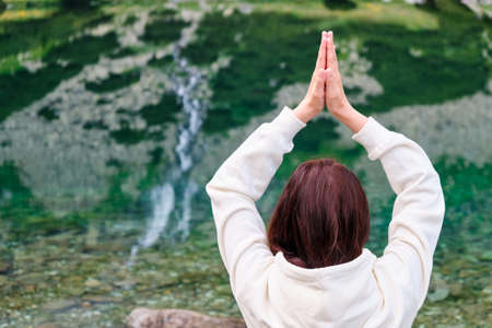 Young woman practicing yoga and meditating on the lake shore with transparent water and reflected...