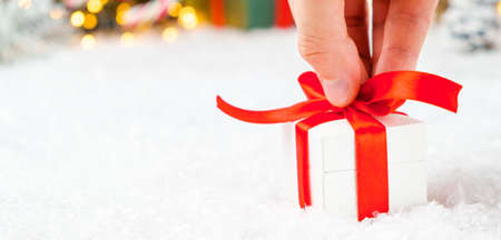 Close up mans hand putting a gift box with a red ribbon on the snow on the background of Christmas...