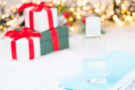Gift box with a red bow from ribbon, sanitizer bottle on the protective face mask against coronavirus on the background of presents under Christmas tree.の写真素材