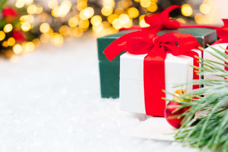 Present with red ribbon, pine or spruce branch with red balls on the snow on the background of blurred Christmas tree and lights of garland.の写真素材