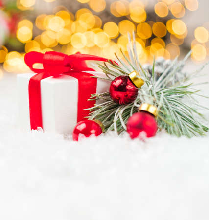Present with red ribbon, pine or spruce branch with red balls on the snow on the background of blurred Christmas tree and lights of garland.の写真素材