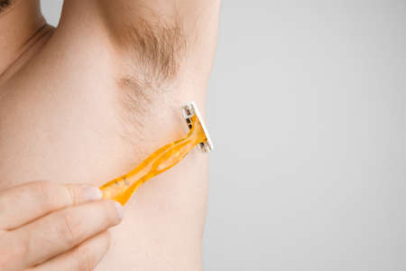 Close up man shaves hairy armpits with a yellow razor. Unshaved armpits or underarm. Depilation and hair remove procedure. Body shaming conceptの写真素材