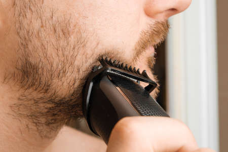 Man shaving beard and cheeks with an electric razor. Morning routine. Trimming beard with a trimmer.