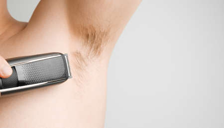 Close up man shaves hairy armpits with an electric razor. Unshaved armpits or underarm. Depilation and hair remove procedure using trimmer. Body shaming conceptの写真素材