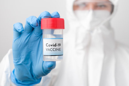 Covid 19 vaccine bottle in doctors hand. Doctor in protective suit, face mask, safety googles and rubber gloves demonstrate vaccine against coronavirus.の写真素材