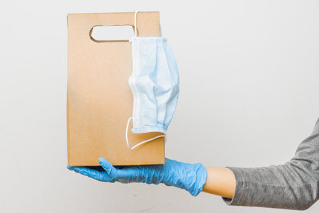 Delivery service concept during quarantine. Woman hand holding a cardboard bag with protective mask on it. Safe delivery from online shops.の写真素材