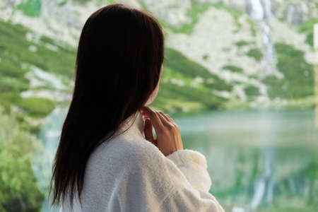 Young woman in a white bathrobe watching in the window with amazing view on the mountains and waterfall. SPA and wellness concept.の写真素材