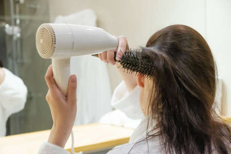 Young woman drying hair with hairdryer and styling hair with round hair brush in the bathroom. Morning routine.の写真素材