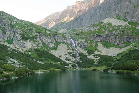 Reflections in the calm lake water, waterfall and mountains at sunset.
