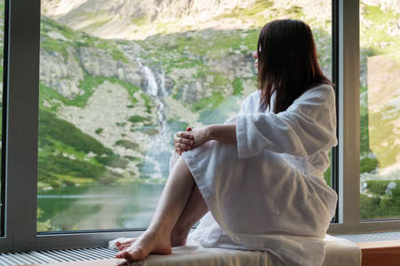 Young woman is sitting on the windowsill and enjoying of amazing view on the mountains, waterfall and lake. SPA and wellness conceptの写真素材