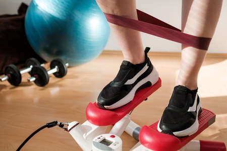 Close woman legs doing cardio on the stepper and using elastic bands with ball, and dumbbells on the background. Workout at home.の写真素材