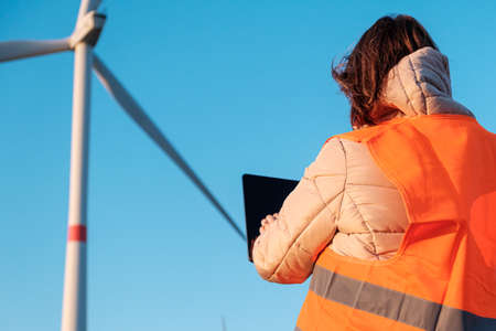 A woman engineer, holding a tablet and working at the wind turbine farm in the field in the orange vestaの写真素材