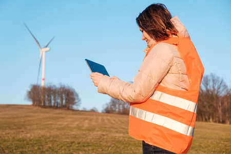 Female engineer checking or repairing wind mills or wind turbines using a tablet in an orange vesta.