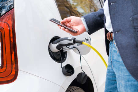 Close up a man in the suit standing near the charging electric car and using his smartphone for...