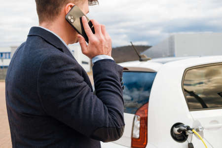 Business man standing near the charging electro and talking on the smartphoneの写真素材