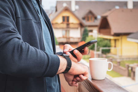 Man using his smartwatch and mobile phone on the balcony and drinking coffee with an amazing view on...