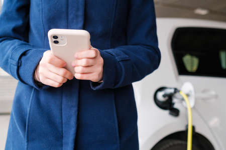 Woman using her mobile phone on the background of the charging electric car at the parking