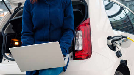Woman sits on the trunk of a car and uses a notebook while the EV is chargingの写真素材
