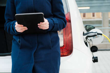 Woman sits on the trunk of a car and uses a tablet while the EV is charging