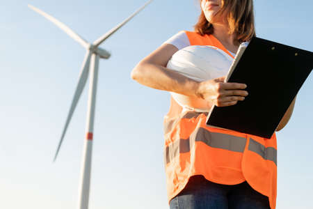 Woman in the orange vest and helmet in the hand holds documents on maintenance of wind turbines on...