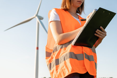 Woman in the orange vest makes notes on maintenance of wind turbines on the background of windmill...