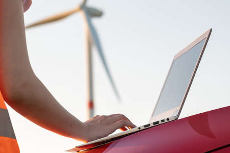 Close up woman hands working on the laptop on the hood of a car against the background of a wind turbine. Maintenance of windmill for generation of renewable and green electricity.の写真素材