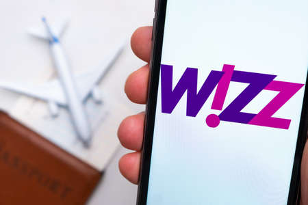 Wizz Airline application on the smartphone screen mans hand. A white toy airplane and a passport are...