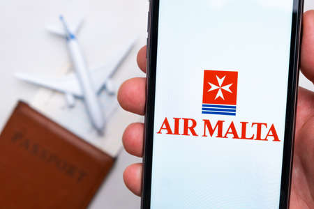 Airmalta Airline application on the smartphone screen mans hand. A white toy airplane and a passport...