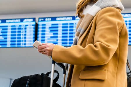 Woman traveler in stylish yellow coat with warm scarf holds mobile phone and suitcase standing...