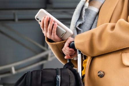 Female passenger in coat with scarf does flight check-in by mobile phone holding large suitcase in...