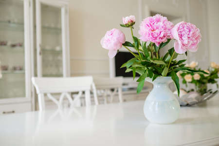 Pink blooming peony flowers with lush green leaves put in vase on white table in light room with modern furniture at home closeupの写真素材
