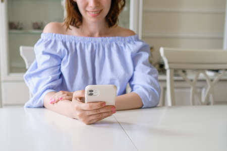 Young happy brunette woman in stylish blouse checks mail in mobile phone sitting at white table in early morning at home close viewの写真素材