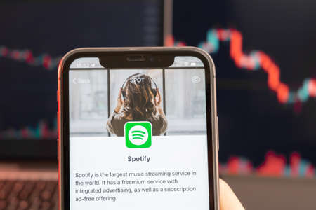 Spotify stock price decrease on the trading market with downtrend line graph bar chart on the background. Man holding a mobile phone with company logo, February 2022, San Francisco, USA.のeditorial素材