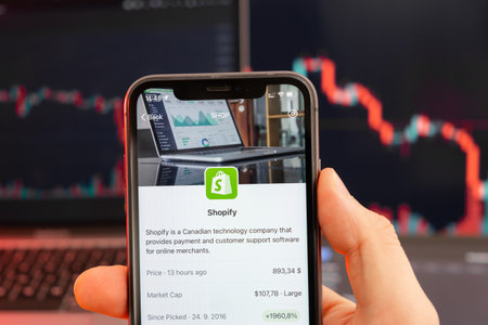 Shopify stock price downtrend with red graphs on the background. Man hands holding a smartphone with...