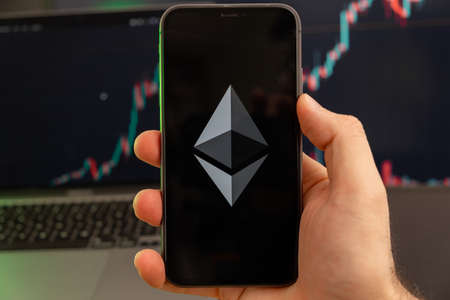 Ethereum ETH app of cryptocurrency stock market analysis on the screen of mobile phone in man hands and growing charts trading data on the background, February 2022, San Francisco, USA.のeditorial素材