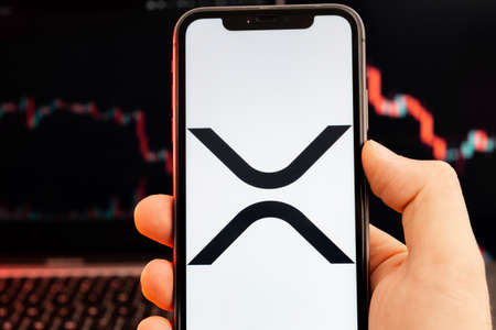 XRP cryptocurrency logo on the screen of smartphone in mans hand with downtrend on the chart on a...